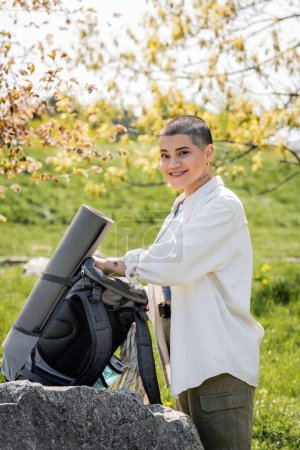 Smiling short haired and tattooed woman hiker in casual clothes looking at camera near backpack on stone with blurred scenic landscape at background , confident female explorer, summer
