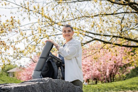 Pleased young short haired and tattooed woman traveler in casual clothes looking away while standing near backpack with travel equipment on stone with nature at background, confident female explorer