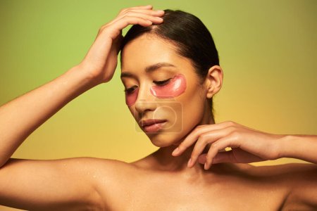 Téléchargez les photos : Campagne de beauté, jeune femme asiatique avec des cheveux bruns et une peau propre posant avec les mains près du visage sur fond vert, épaules nues, patchs oculaires hydratants, peau éclatante - en image libre de droit