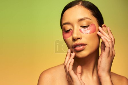 skincare campaign, sensual asian woman with brunette hair and clean skin posing with hands near face on green background, bare shoulders, moisturizing eye patches, glowing skin 