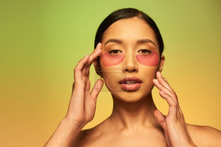 beauty product, young asian woman with brunette hair and clean skin posing and looking at camera on green background, bare shoulders, moisturizing eye patches, glowing skin 