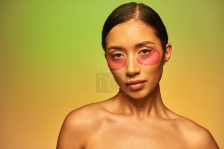 skincare campaign, young asian woman with brunette hair and bare shoulders posing and looking at camera on green background, face care, moisturizing eye patches, glowing skin 