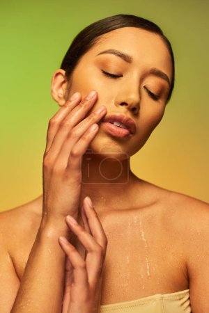 Flüssigkeitszufuhr, junge asiatische Frau mit nackten Schultern und nassem Körper posiert auf Gradientenhintergrund, geschlossene Augen, anrührende Wange, Hautpflege-Kampagne, Schönheitsmodel, brünettes Haar, glühende Haut 