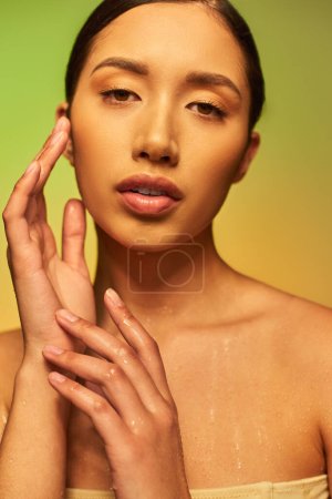 Flüssigkeitszufuhr, junge asiatische Frau mit nackten Schultern und nassem Körper posiert auf Gradienten Hintergrund, berühren Gesicht, Blick in die Kamera, Hautpflege-Kampagne, Beauty-Modell, brünettes Haar, glühende Haut 