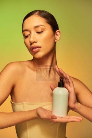 Produktpräsentation, Hautpflegeprodukt, junge asiatische Frau mit nackten Schultern hält Kosmetikflasche mit Körperlotion und posiert auf grünem Hintergrund, glühende Haut, brünettes Haar 