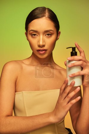 product presentation, skin care product, young asian woman with bare shoulders holding cosmetic bottle with body lotion and posing on green background, glowing skin concept 