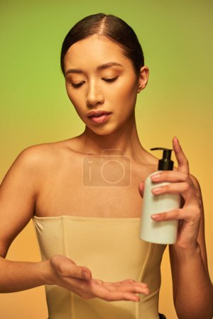 product presentation, skin care product, young asian woman with bare shoulders holding cosmetic bottle and posing on green background, glowing skin, brunette hair 