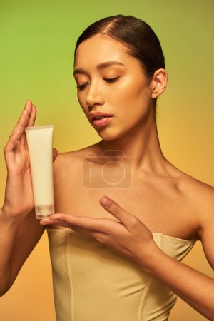 skin care, product presentation, youthful skin, young asian woman with bare shoulders holding cosmetic tube and posing on green background, glowing skin, brunette hair 