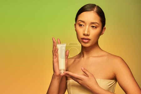 Schönheit und Hautpflege, Produktpräsentation, junge asiatische Frau mit nackten Schultern hält Kosmetikschlauch mit Creme und posiert auf Gradienten Hintergrund, glühende Haut, brünettes Haar 