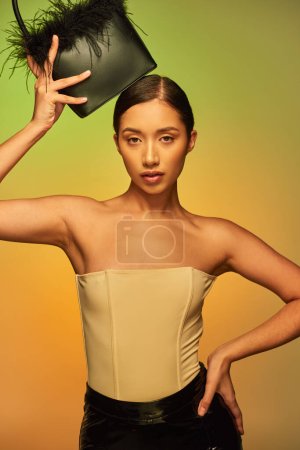 choix de mode, femme asiatique brune aux épaules nues posant avec sac à main en plumes et la main sur la hanche sur fond vert, dégradé, déclaration de mode, peau éclatante, beauté naturelle, jeune modèle 