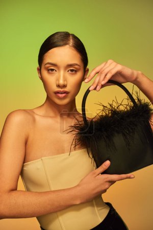 mode avant, femme asiatique brune avec épaules nues posant avec sac à main en plumes sur fond vert, dégradé, déclaration de mode, peau éclatante, beauté naturelle, jeune modèle regardant la caméra