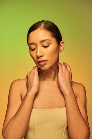 Schönheitsfotografie, asiatische Frau mit brünetten Haaren und nackten Schultern posiert auf Gradienten Hintergrund, grün und orange, Hände in Nackennähe, Hautpflege, glühende Haut, natürliche Schönheit, junges Modell 