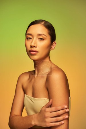 beauté asiatique, jeune femme aux cheveux bruns et épaules nues posant sur fond dégradé, vert et orange, soin de la peau, peau éclatante, beauté naturelle, modèle beauté 