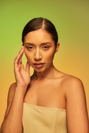 Schönheitsfotografie, asiatische Frau mit brünetten Haaren und nackten Schultern posiert auf Gradienten Hintergrund, Hautpflege, glühende Haut, natürliche Schönheit, junges Modell posiert auf grünem und orangefarbenem Hintergrund 