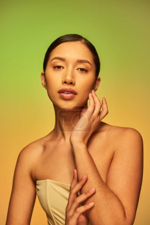 Schönheitsfotografie, hübsche asiatische Frau mit brünetten Haaren und nackten Schultern posiert auf Gradienten Hintergrund, grün und orange, Hautpflege, glühende Haut, natürliche Schönheit, junges Modell 