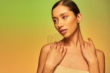 Schönheits- und Hautpflege, hübsche asiatische Frau mit brünetten Haaren und nackten Schultern posiert auf Gradientenhintergrund, grün und orange, Hautpflege, glühende Haut, natürliche Schönheit, junges Model 
