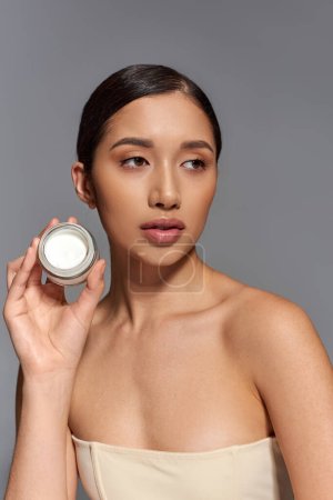 Produktpräsentation, Hautpflege, junges asiatisches Model mit brünetten Haaren, Schönheitsglas mit Creme auf grauem Hintergrund, glühende und heide Haut, Schönheitskampagne, Gesichtsbehandlungskonzept 