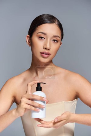 campagne de beauté, présentation de produits, soins de la peau, jeune modèle asiatique avec cheveux bruns tenant bouteille cosmétique sur fond gris, peau éclatante et paresseuse, concept de traitement du visage  