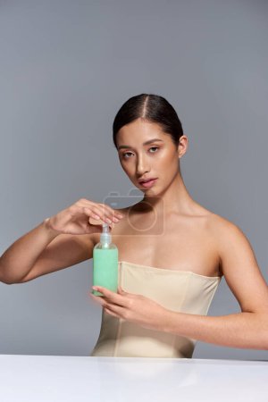 Produktpräsentation, Hautpflege, junges asiatisches Model mit brünetten Haaren mit Kosmetikflasche auf grauem Hintergrund, glühende und heide Haut, Schönheitskampagne, Gesichtsbehandlungskonzept 