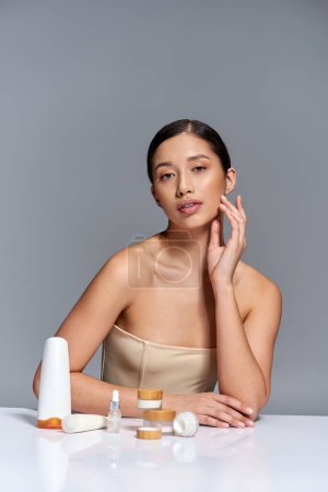 Hautpflege-Präsentation, junges asiatisches Model mit brünetten Haaren posiert in der Nähe verschiedener Schönheitsprodukte auf grauem Hintergrund, glühende und heide Haut, Schönheitskampagne, Gesichtsbehandlungskonzept 