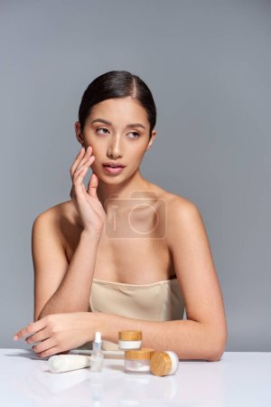 présentation de soins de la peau, jeune femme asiatique aux épaules nues posant à proximité de différents produits de beauté sur fond gris, peau luisante et paresseuse, campagne de beauté, concept de traitement du visage 