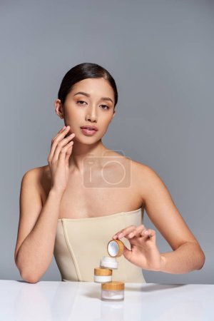 Produktpräsentation, Hautpflege, junges asiatisches Model mit brünetten Haaren, die Gesichtscreme auf grauem Hintergrund halten, glühende und heide Haut, Schönheitskampagne, Gesichtsbehandlung, konzeptionelle 