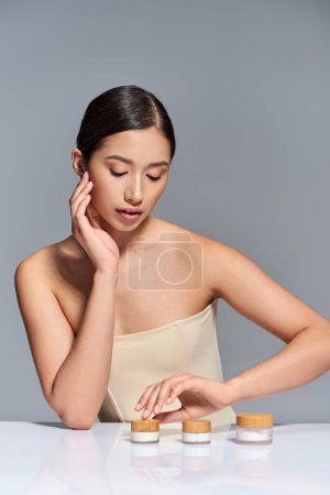 Hautpflege, junges asiatisches Model mit brünetten Haaren posiert in der Nähe verschiedener Schönheitsprodukte auf grauem Hintergrund, glühende und heide Haut, Schönheitskampagne, Gesichtsbehandlungskonzept 