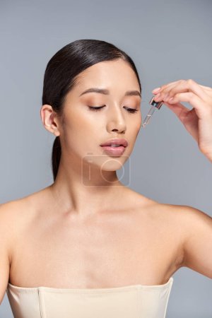 skin care, young asian woman with brunette hair applying serum with pipette on grey background, glowing and heathy skin, beauty campaign, facial treatment concept 