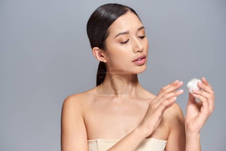 Produktpräsentation, Hautpflege, junges asiatisches Model mit brünetten Haaren im Kosmetikglas mit Creme auf grauem Hintergrund, glühende und heide Haut, Schönheitskampagne, Gesichtsbehandlungskonzept 