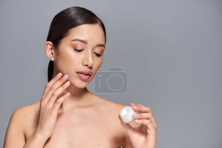 skin care, young asian model with brunette hair holding cosmetic jar and applying face cream on grey background, glowing and heathy skin, beauty campaign, facial treatment concept 