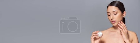 Photo for Skin care, young asian woman with brunette hair holding cosmetic jar and applying face cream on grey background, glowing and heathy skin, beauty campaign, facial treatment concept, banner - Royalty Free Image