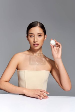 product presentation, skin care, young asian model with brunette hair holding cosmetic jar with eye cream on grey background, glowing and heathy skin, beauty campaign, facial treatment concept 