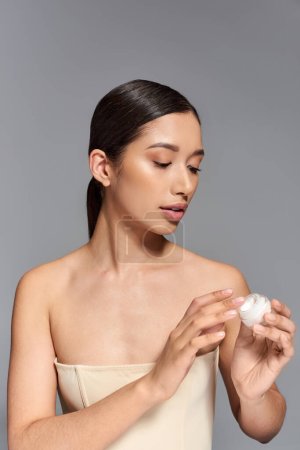 beauty campaign, skin care, young asian woman with brunette hair holding cosmetic jar with eye cream on grey background, glowing and heathy skin,  facial treatment, beauty industry 