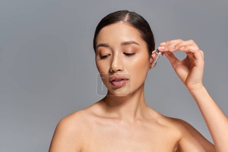 beauty campaign, skin care, young asian woman with brunette hair applying serum with pipette on grey background, glowing and heathy skin, facial treatment concept, looking away