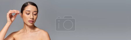 Téléchargez les photos : Campagne de beauté, soin de la peau, jeune femme asiatique avec cheveux bruns appliquant sérum avec pipette sur fond gris, peau éclatante et paresseuse, concept de traitement du visage, détournement des yeux, bannière - en image libre de droit