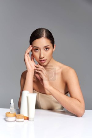 Schönheitsindustrie, Hautpflege, junge asiatische Frau mit brünetten Haaren posiert in der Nähe von Schönheitsprodukten auf grauem Hintergrund, glühende und heide Haut, Gesichtsbehandlungskonzept, Gesichtspflege 