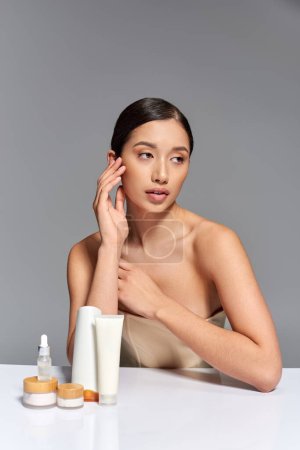 Schönheitstrend, Hautpflege, junge asiatische Frau mit brünetten Haaren posiert in der Nähe von Schönheitsprodukten auf grauem Hintergrund, glühende und heide Haut, Gesichtsbehandlungskonzept, Gesichtspflege 