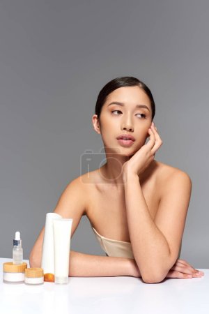 beauty industry, young asian woman with brunette hair posing near beauty products on grey background, glowing and heathy skin, facial treatment concept, facial and skin care 
