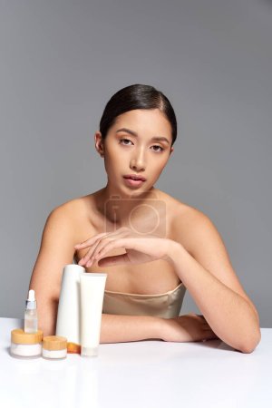 photographie de beauté, jeune femme asiatique aux cheveux bruns posant des produits de beauté proches sur fond gris, peau éclatante et païenne, concept de traitement du visage, soins du visage et de la peau, jeunesse 