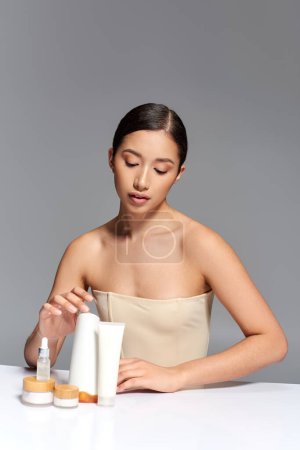 facial treatment, young asian woman with brunette hair posing near beauty products on grey background, glowing and heathy skin, facial care concept, asian youth, beauty products