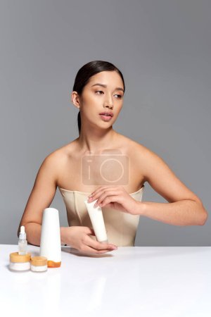 beauty campaign, young asian woman with brunette hair posing near beauty products on grey background, glowing and heathy skin, facial treatment concept, face and skin care, youth 