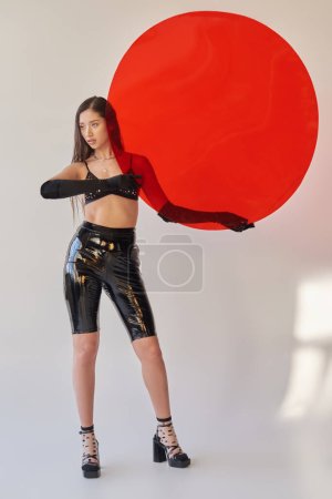 belleza y moda, estilo látex, mujer asiática joven en sujetador y guantes sosteniendo vidrio rojo en forma redonda sobre fondo gris, opciones de moda, cabello moreno, traje elegante, juventud 