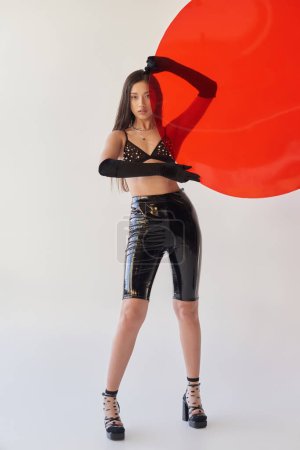 beauty and fashion, latex style, young asian woman in bra and gloves holding red round shaped glass on grey background, fashion statement, brunette hair, stylish outfit, youth 