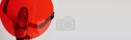 Téléchargez les photos : Photographie de mode, style latex, jeune femme asiatique avec des cheveux bruns posant en soutien-gorge et gants et tenant verre rond rouge sur fond gris, choix de mode, derrière le verre, bannière - en image libre de droit