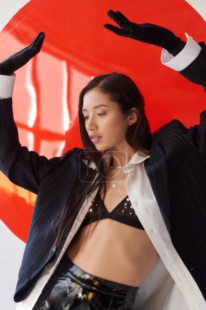 edgy fashion, young asian woman in bra, white shirt and blazer posing in gloves near red round shaped glass, grey background, personal style, underwear and jacket, youth 