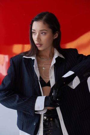 edgy fashion, young asian woman in bra, white shirt and blazer posing in gloves and latex pants near red round shaped glass, grey background, personal style, underwear and jacket, youth 