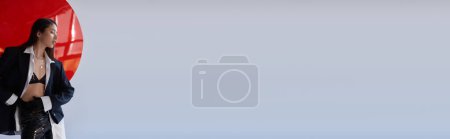 moda vanguardista, mujer asiática joven en sujetador, camisa blanca y chaqueta posando en pantalones de látex y guantes negros y pantalones de látex cerca de vidrio redondo rojo, fondo gris, ropa interior y chaqueta, pancarta 