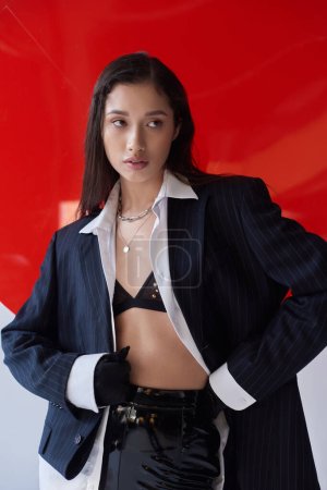 Photo for Stylish look, young asian woman in bra, white shirt and blazer posing in gloves near red round shaped glass, looking away on grey background, personal style, underwear and jacket, youth - Royalty Free Image