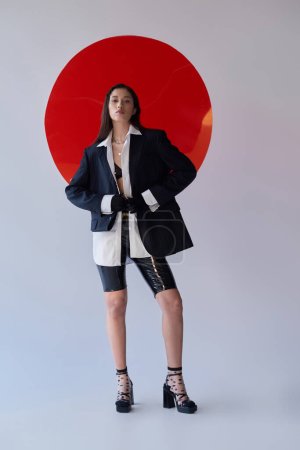 stylish look, young asian woman in bra, white shirt and blazer posing in gloves and latex shorts near red round shaped glass, looking away on grey background, personal style, youth, full length 
