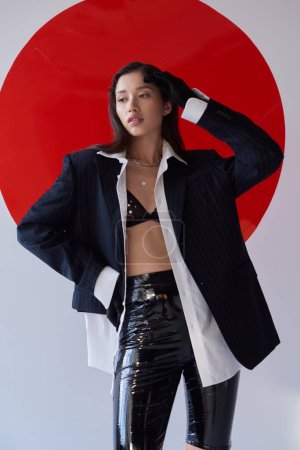 stylish outfit, young asian woman in bra, white shirt and blazer posing in gloves and latex shorts near red round shaped glass, looking away on grey background, personal style, youth 
